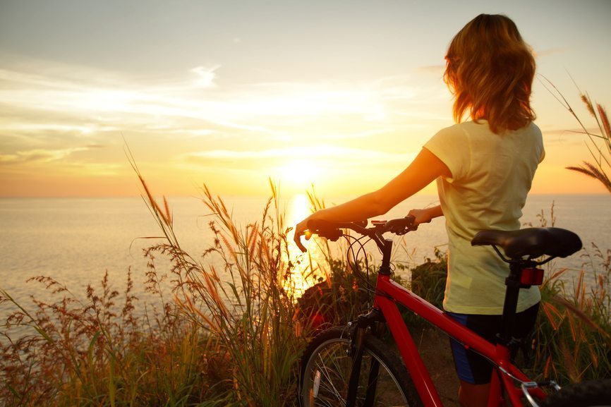 Young lady standing with bicycle on a sea coast and enjoying sunset